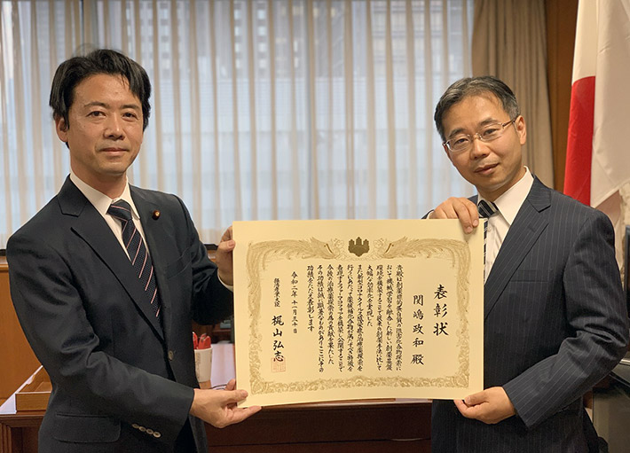 Associate Professor Sekijima receives a certificate of commendation from Parliamentary Vice-Minister Koichi Munekiyo (left) of the Ministry of Economy, Trade and Industry