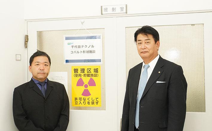 コバルト照射施設の前に立つオープンファシリティセンター安全管理・放射線部門の依田功部門長（左）と井上社長（右）
